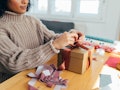 A young Black woman wraps up a present in her home with purple and rust ribbon while wearing a chunk...
