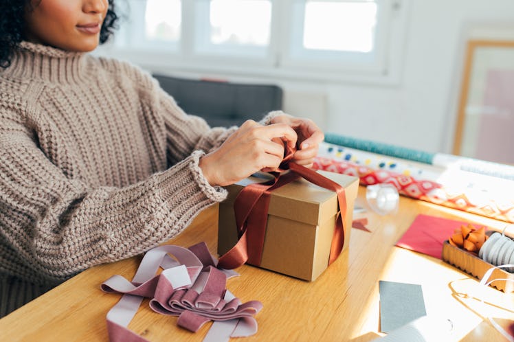 A young Black woman wraps up a present in her home with purple and rust ribbon while wearing a chunk...