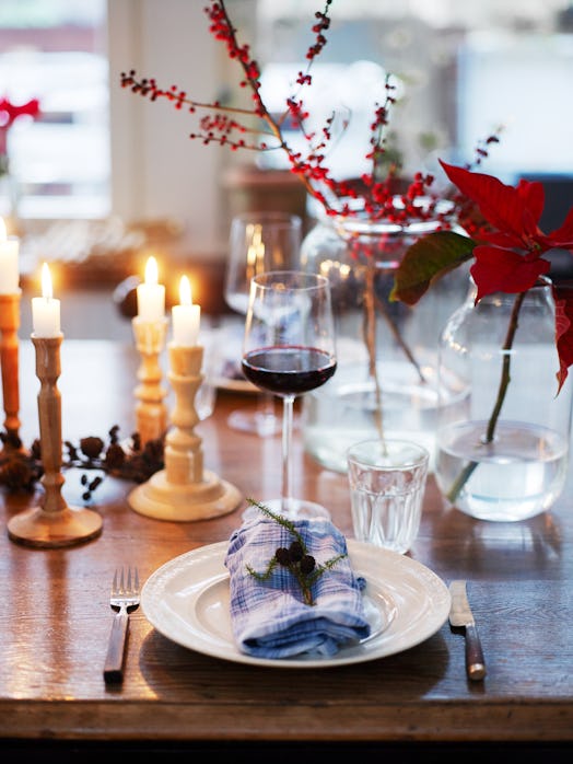 A tablescape featuring candles, red plants and pine tree leaves as well as checkered napkins on plat...