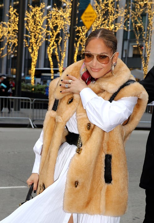 Jennifer Lopez with sunglasses on, wearing a white dress and a fur vest while walking on the street