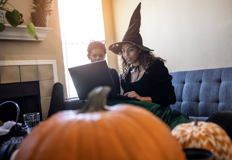 Two young women sit with their laptop on a couch while wearing Halloween costumes and video chatting...