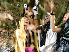 Three women raise Halloween cocktails in the air while dressed up in costumes in the woods.