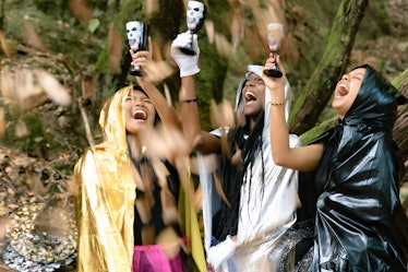Three women raise Halloween cocktails in the air while dressed up in costumes in the woods.