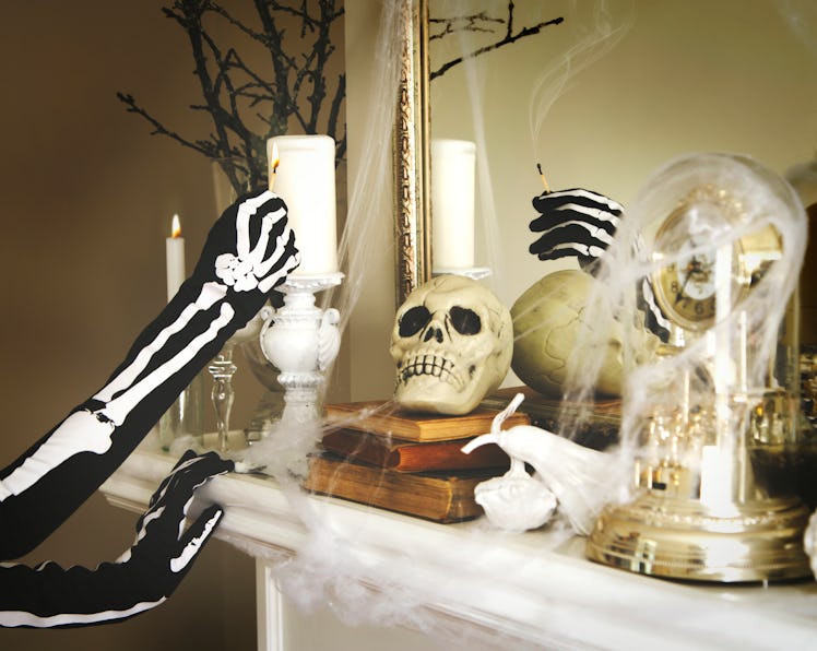 A young woman wearing a skeleton costume lights a candle on a fireplace with Halloween decor.