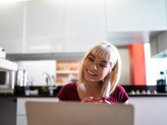 A young Asian woman video chats on her laptop while sitting in her home with a cup of tea.