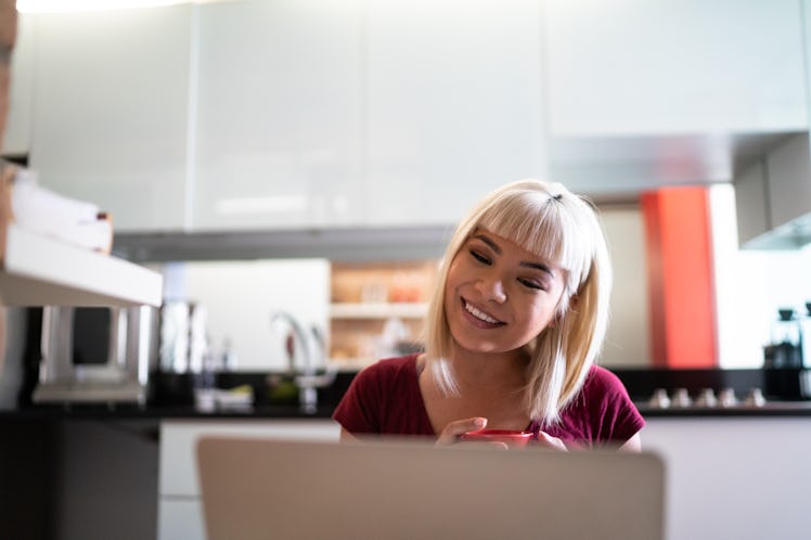 A young Asian woman video chats on her laptop while sitting in her home with a cup of tea.