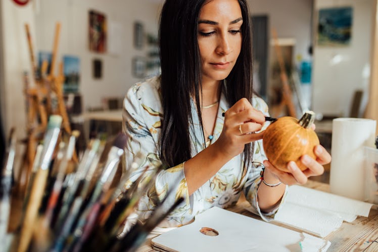 A young woman draws on a pumpkin while sitting at a table in her home.
