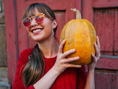 A young woman holds a pumpkin while standing against a red barn and wearing red-tinted sunglasses.