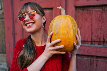 A young woman holds a pumpkin while standing against a red barn and wearing red-tinted sunglasses.