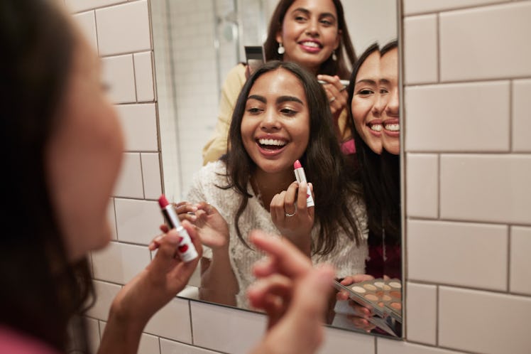 Three friends crowd around a bathroom mirror to apply lipstick. 