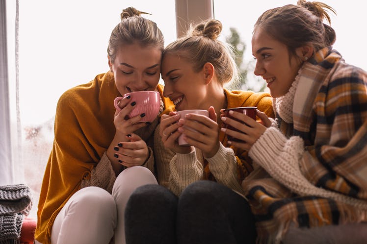 Three girls bundled up for fall hold mugs while sitting in front of a bright window.
