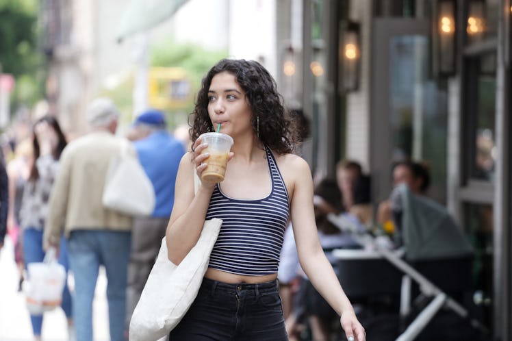 A woman with curly hair wearing a striped tank and black jeans sips her iced coffee while walking do...