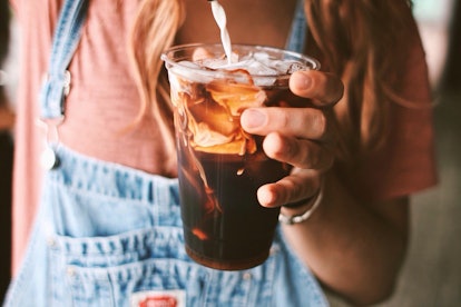 A woman wearing overalls, pours some milk into her cold brew drink. 