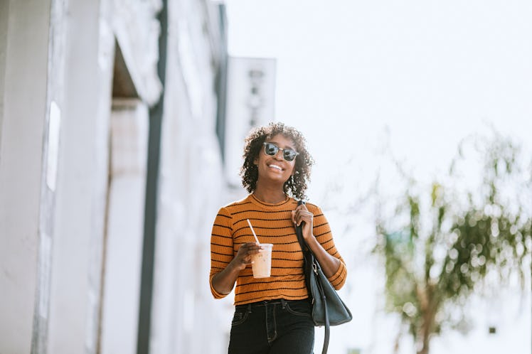 A happy woman in an orange shirt walks down the street with an iced coffee. 
