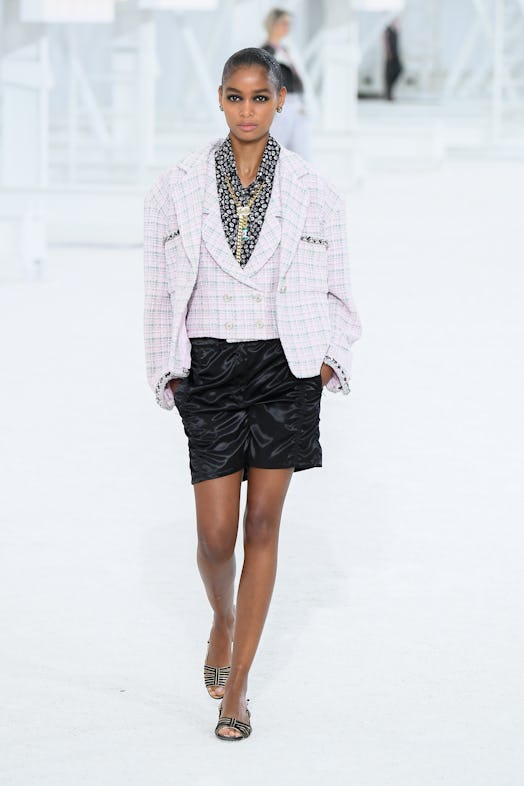 The model wears an iconic Chanel light-pink tweed blazer with a matching vest paired with black shor...
