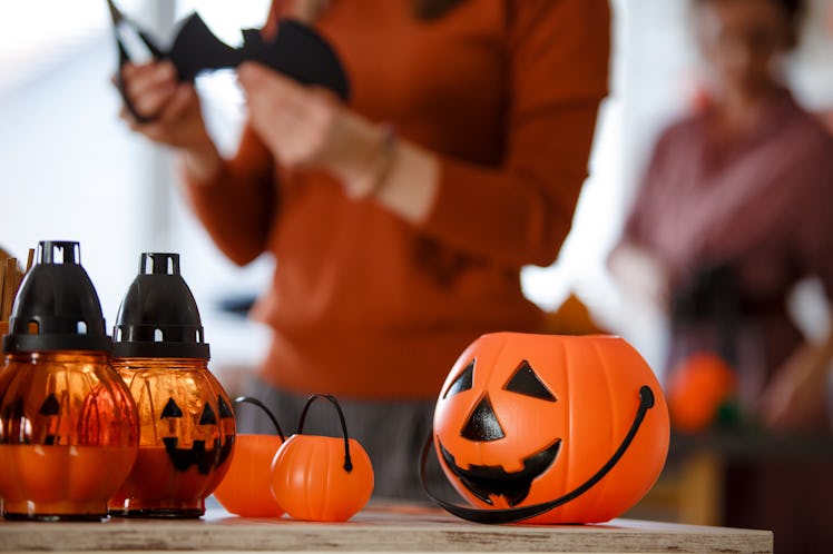 A pumpkin bucket sits on the table, as a woman cuts a bat decoration in the kitchen.