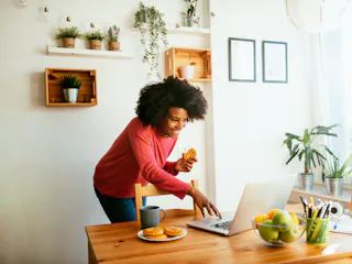 A young Black woman video chats with her friends while eating a dessert in her kitchen.