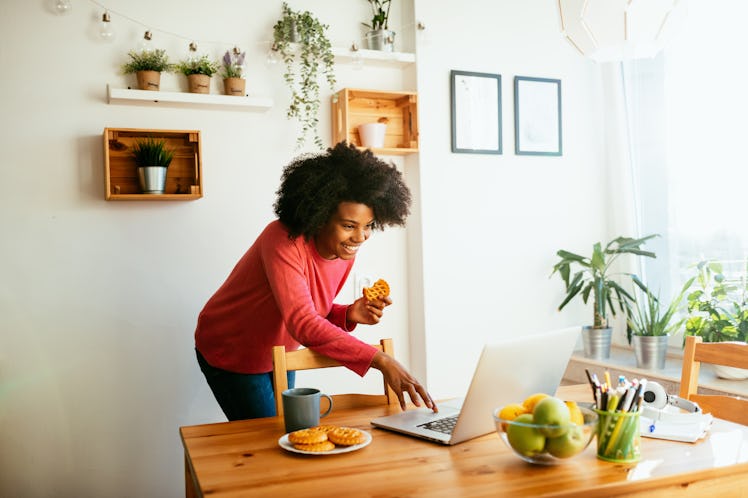 A young Black woman video chats with her friends while eating a dessert in her kitchen.