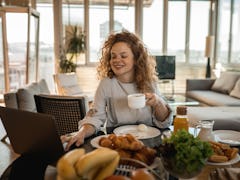 A young woman with curly red hair sits at a table with her laptop and video chats with friends while...