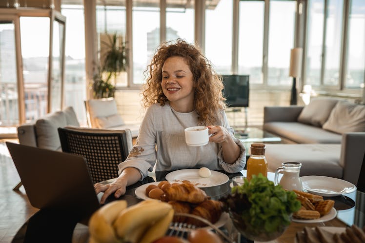 A young woman with curly red hair sits at a table with her laptop and video chats with friends while...