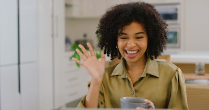 A young Black woman waves at her laptop while video chatting with friends and drinking coffee.