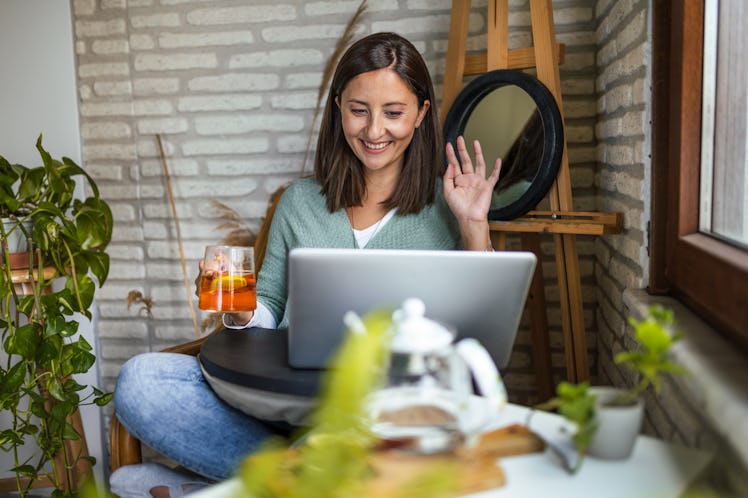 A young woman waves to friends she's video chatting with while drinking tea and sitting near plants.