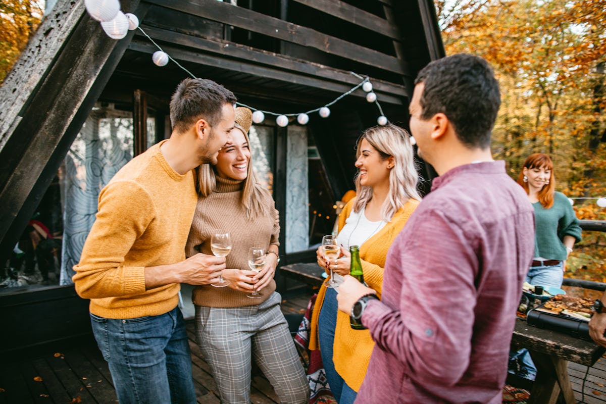 Two trendy couples smile and enjoy drinks on the deck of a cabin for a Friendsgiving gathering.