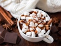 A cup of hot chocolate with marshmallows sits on a wooden table with chunks of chocolate, cinnamon s...