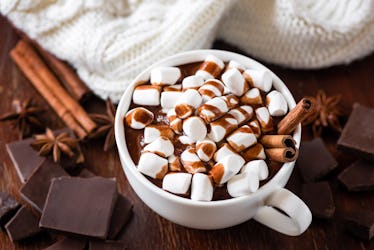 A cup of hot chocolate with marshmallows sits on a wooden table with chunks of chocolate, cinnamon s...