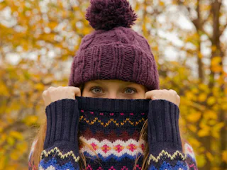 A woman wearing a beanie holds up her sweater over her mouth, while standing outside in the fall.
