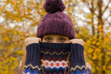 A woman wearing a beanie holds up her sweater over her mouth, while standing outside in the fall. 
