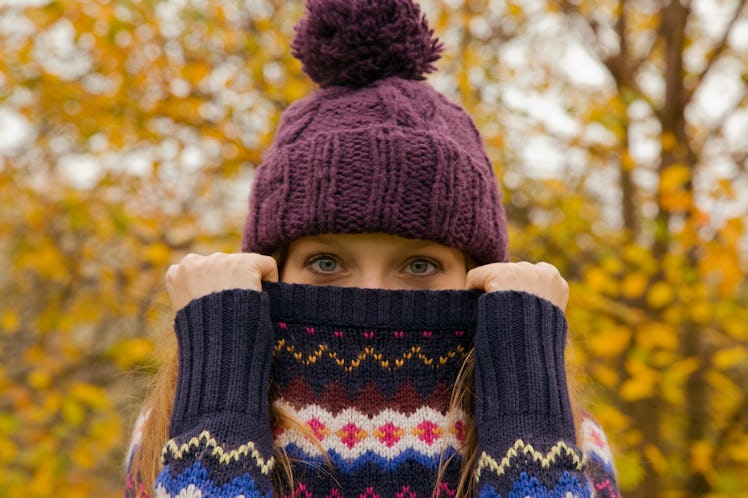 A woman wearing a beanie holds up her sweater over her mouth, while standing outside in the fall.