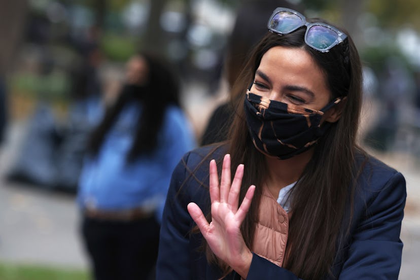 Alexandria Ocasio-Cortez waving while wearing a black jacket and a black face mask