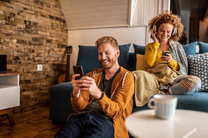 A happy couple relaxes at home while looking at their phones in a trendy living room.