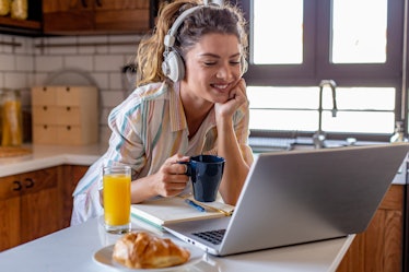 A happy woman with headphones and a striped button-down shirt on holds a coffee mug while smiling at...