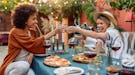 Two trendy friends laugh and toast their wine glasses while enjoying a Friendsgiving meal outside.