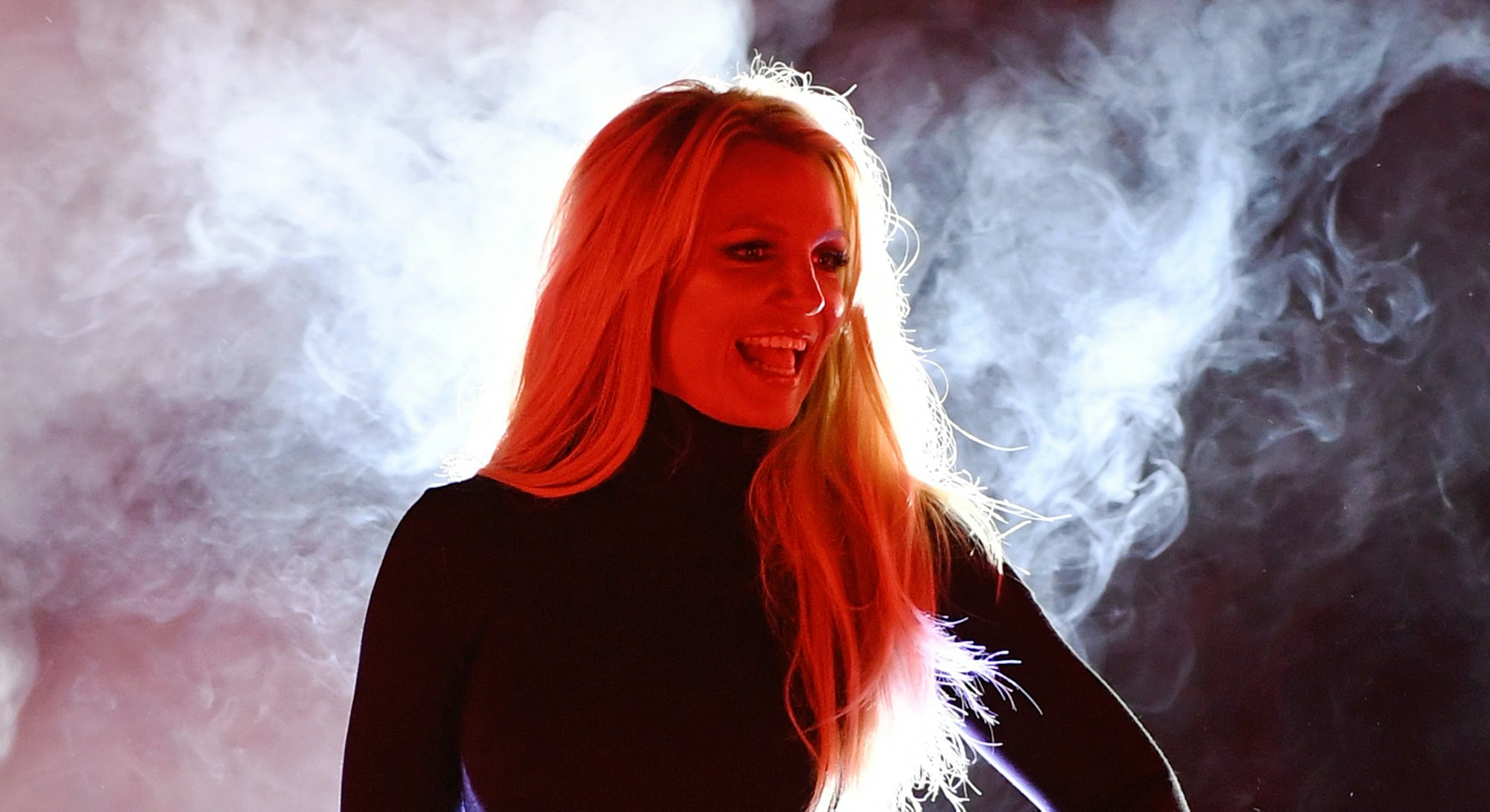 Britney Spears wearing a black dress with two golden starts and smiling while standing on the stage 
