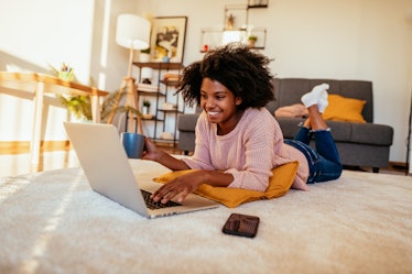 A happy Black woman holds a coffee mug while typing on her computer laying on the ground.