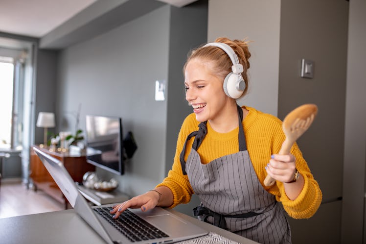 A happy woman holding a wooden spoon checks her laptop while cooking in the kitchen.