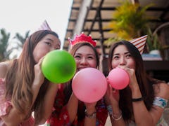 Three friends, wearing party hats, blow up balloons at a birthday party. 