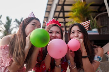 Three friends, wearing party hats, blow up balloons at a birthday party. 