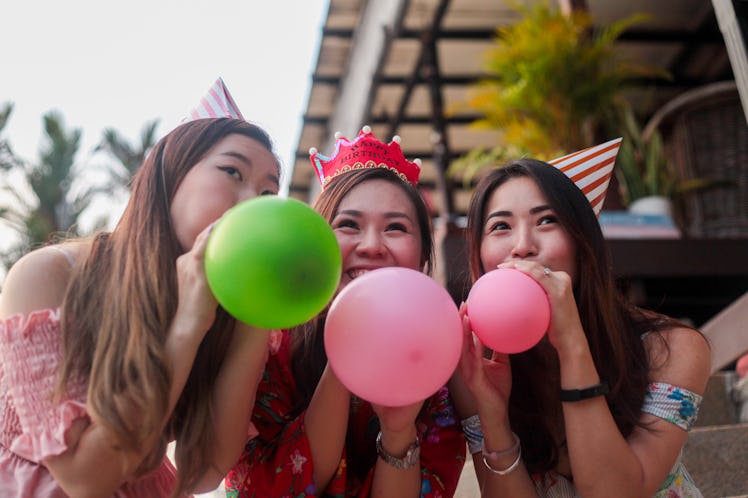 Three friends, wearing party hats, blow up balloons at a birthday party.