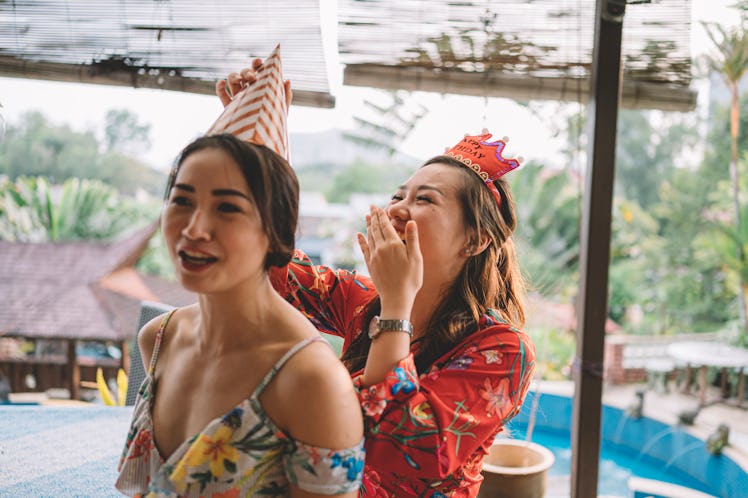 A girl places a birthday hat on her friend, while laughing. 