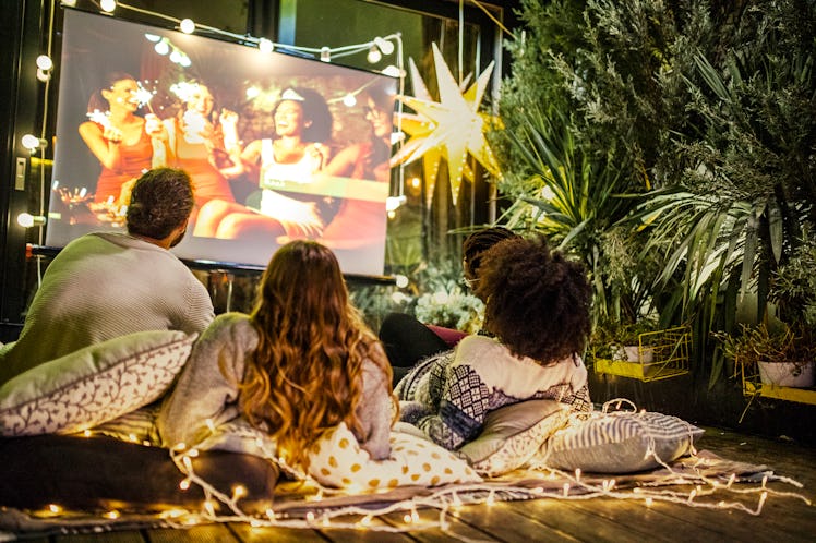 A group of friends watches a movie while sitting on pillows and blankets in their backyard movie the...