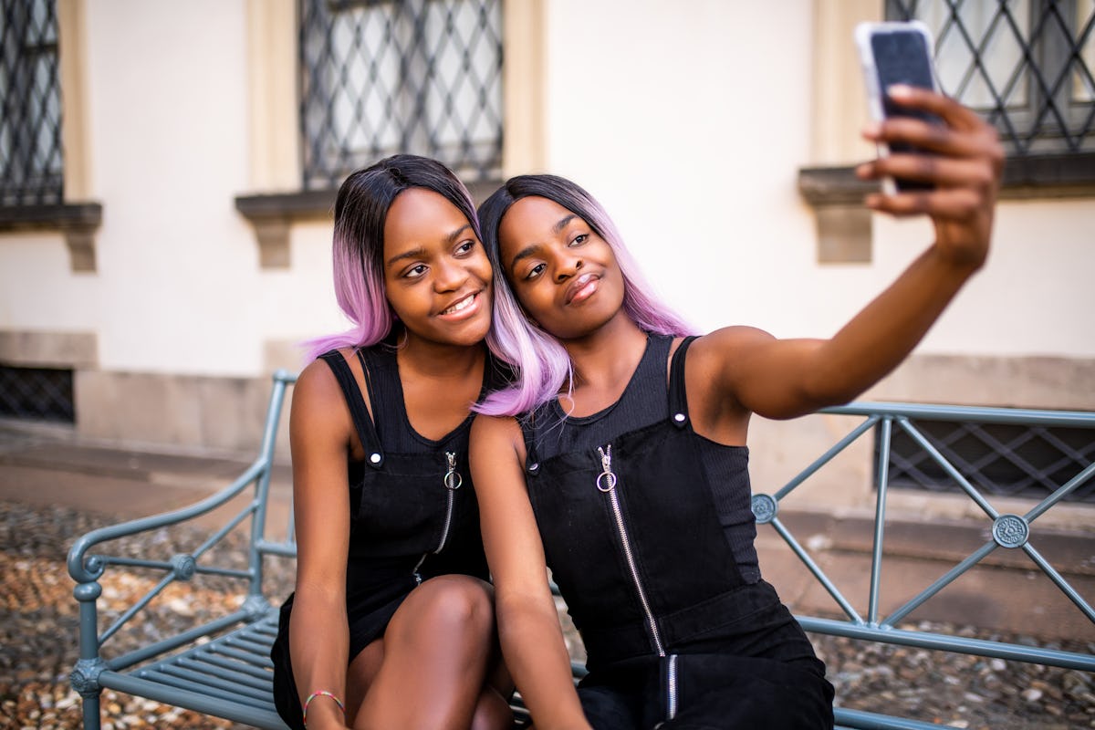 Two twins with black and pink hair pose for a selfie on a bench outside.