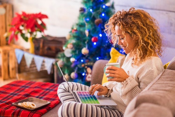 A woman wearing loungewear with a plate of cookies and mug in hand, sits by the Christmas tree, look...