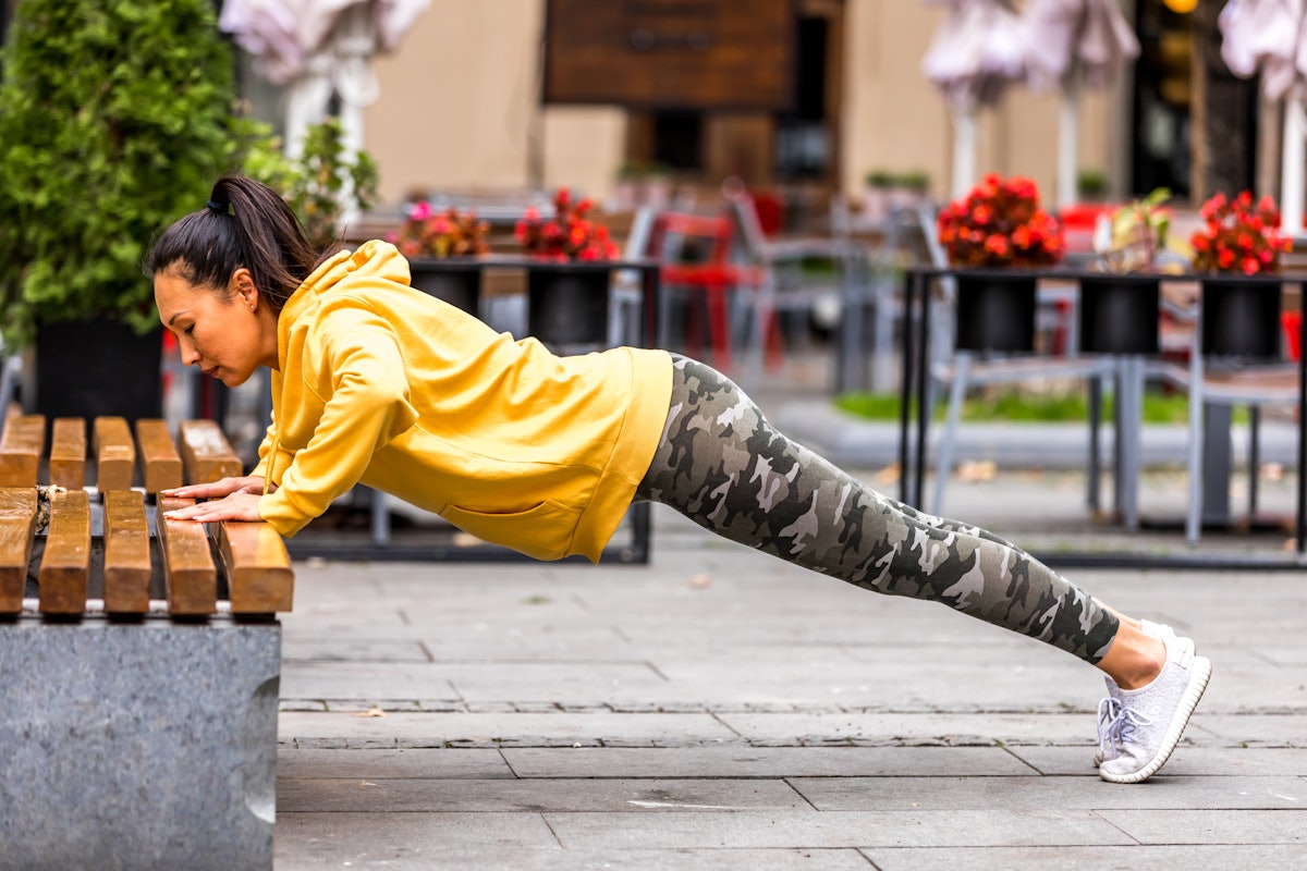 A person wearing leggings and a yellow jacket performs an elevated pushup on a bench. Elevated pushu...