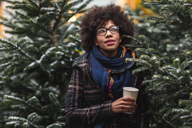 A young Black woman stands amongst a row of evergreen trees and holds a coffee cup.
