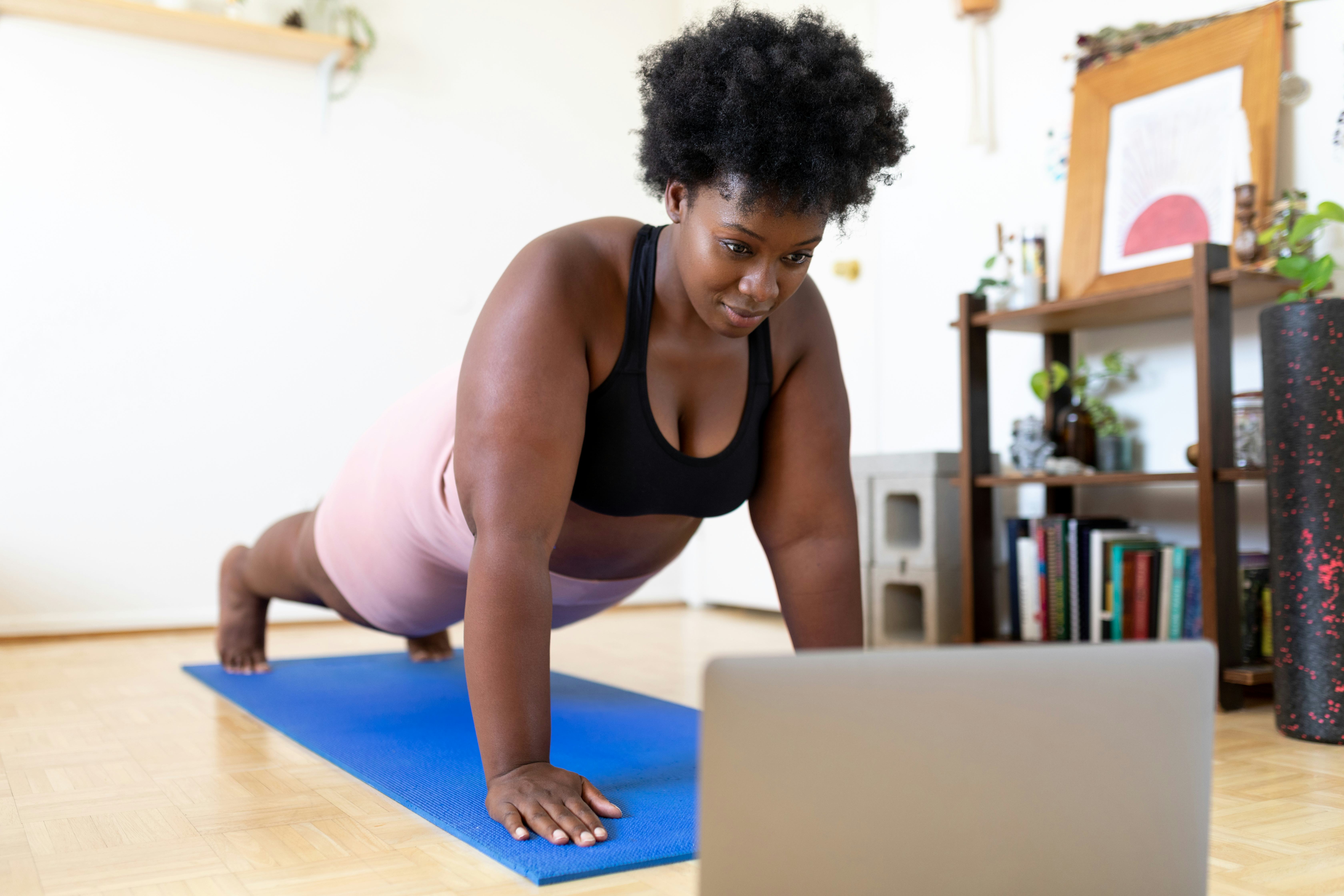 A person with natural hair holds a plank position while looking at their laptop in their living room...