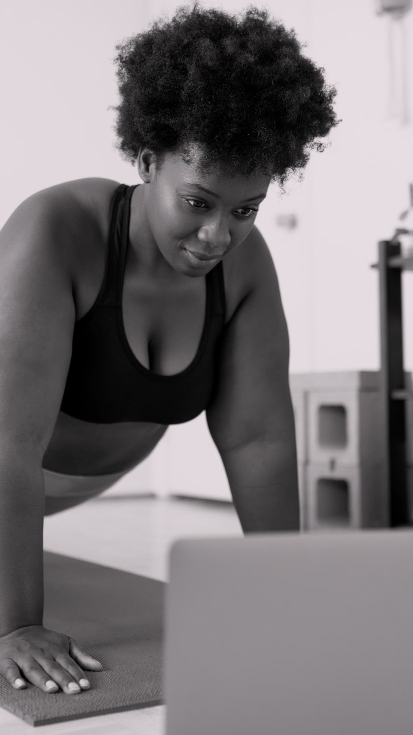 A person with natural hair holds a plank position while looking at their laptop in their living room...